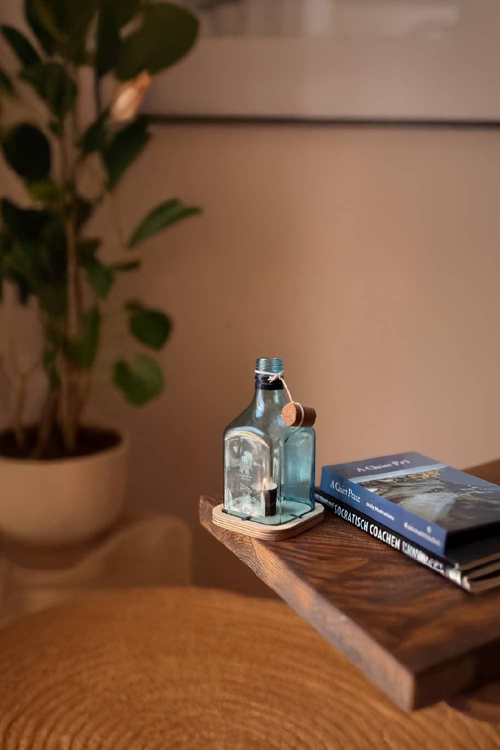 A pale blue glass bottle candle on a round wooden base, with a cork tied to its neck, on a shelf beside books and a plant