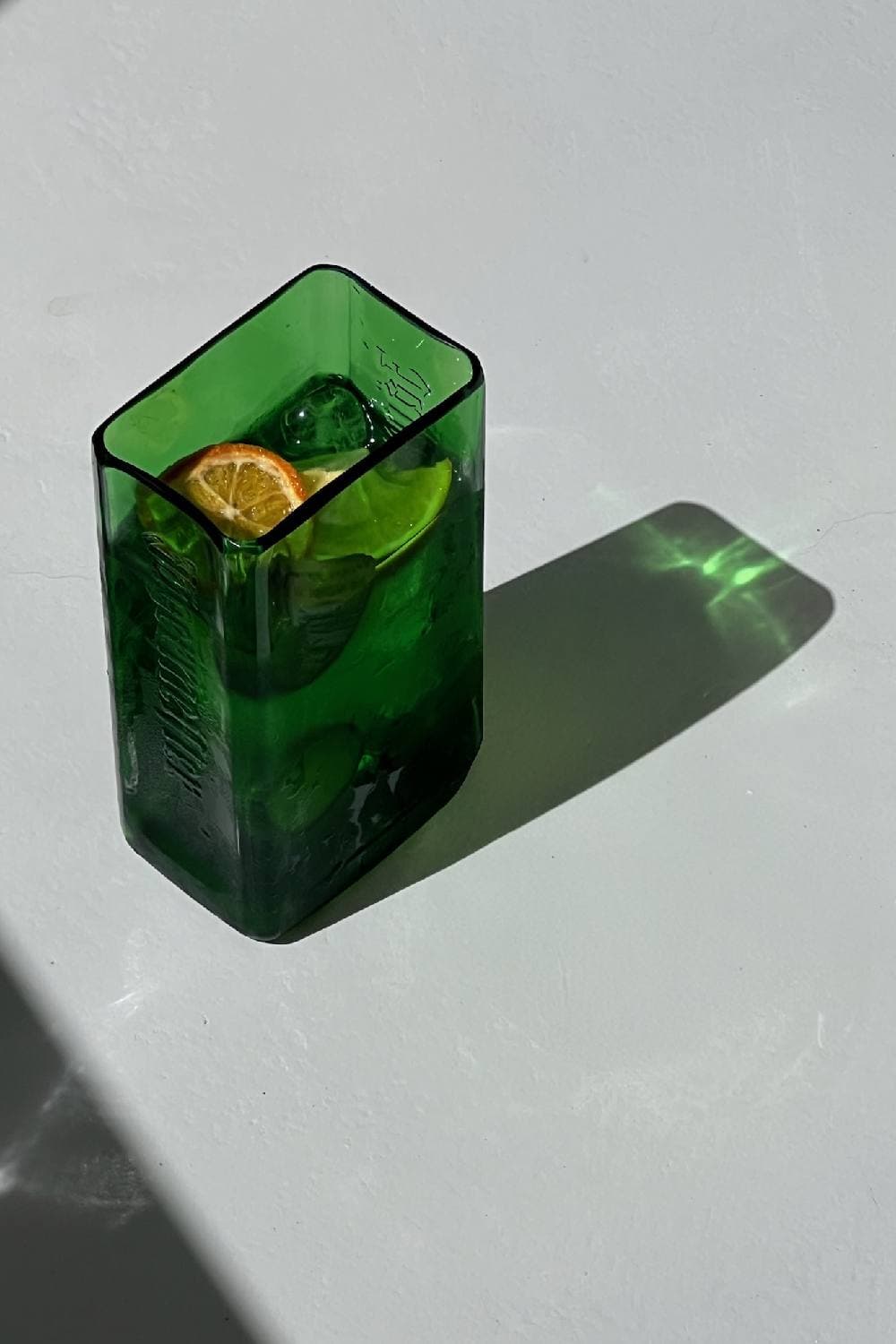 A green glass jug with square shoulders holding ice, a lime wheel and a dried orange slice, casting a green shadow on a white surface