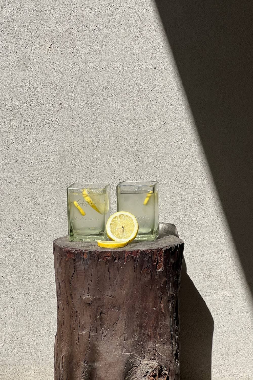 Two clear square glasses with lemon slices on a tree stump against a pale wall, half a lemon beside them