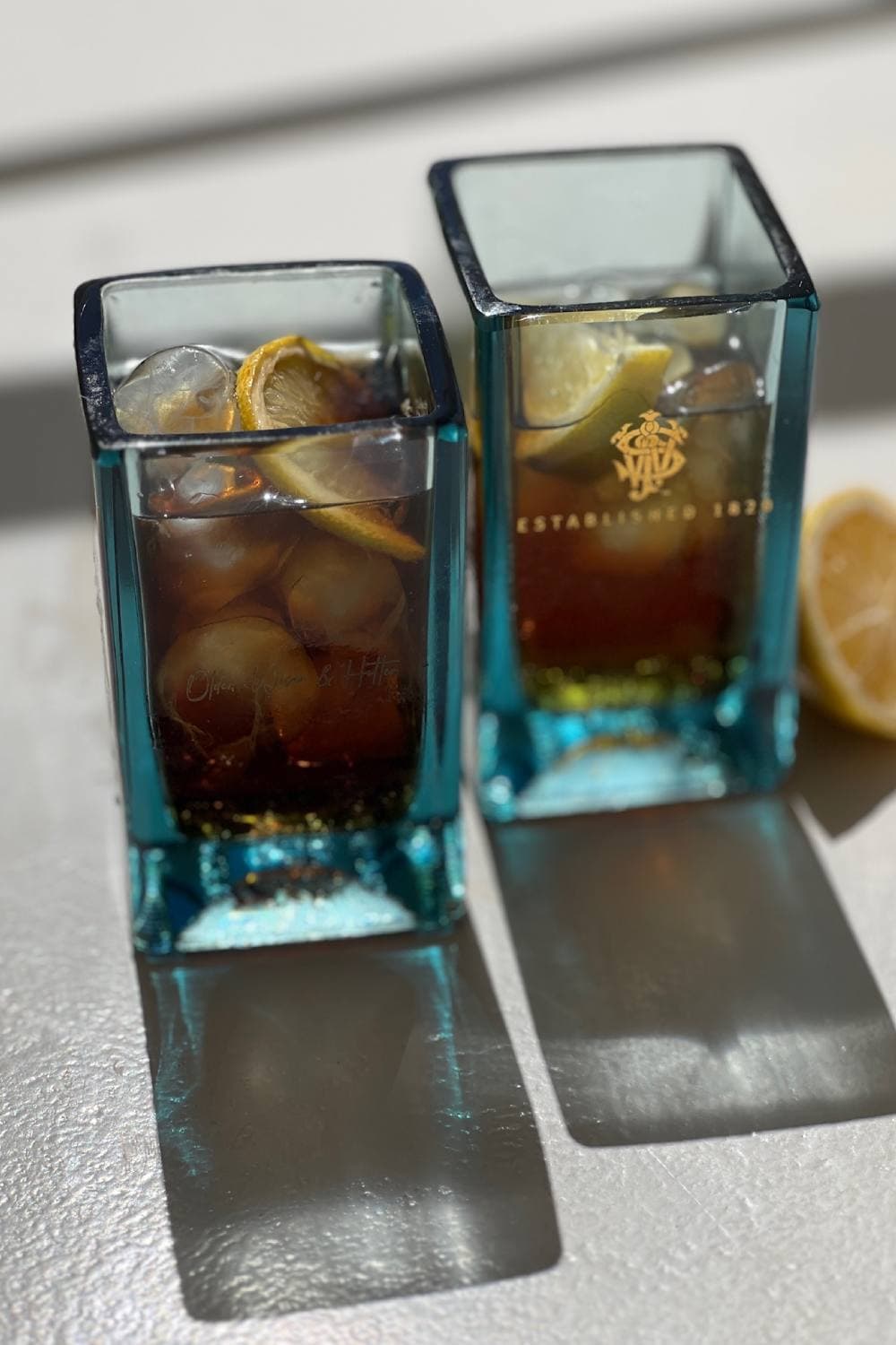 Two sea-blue square glasses of iced drink with lemon, one with a gold crest, on a pale surface in strong sunlight