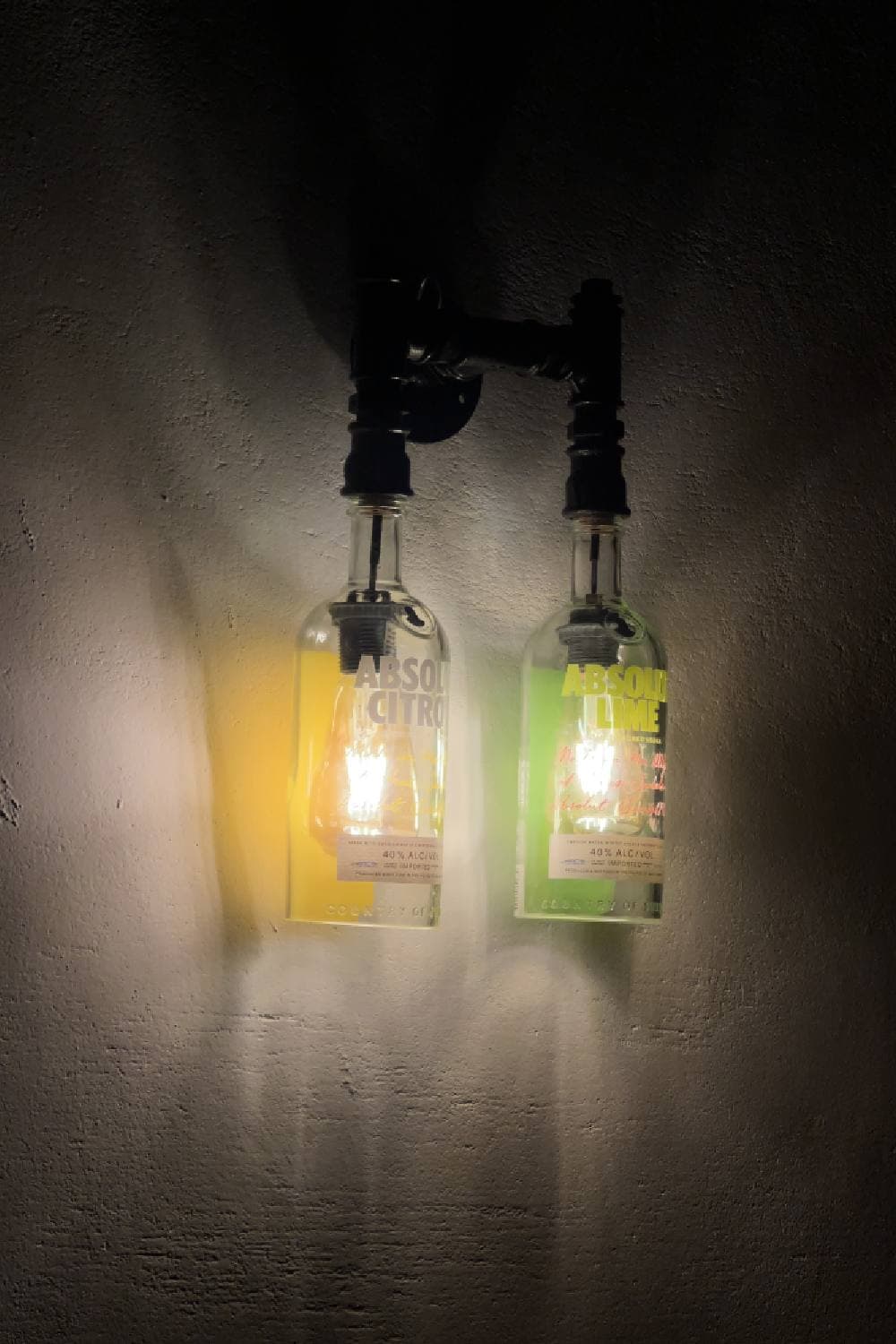 Two bottle wall lights on black pipe fittings, one glowing yellow and one green, against a rough wall