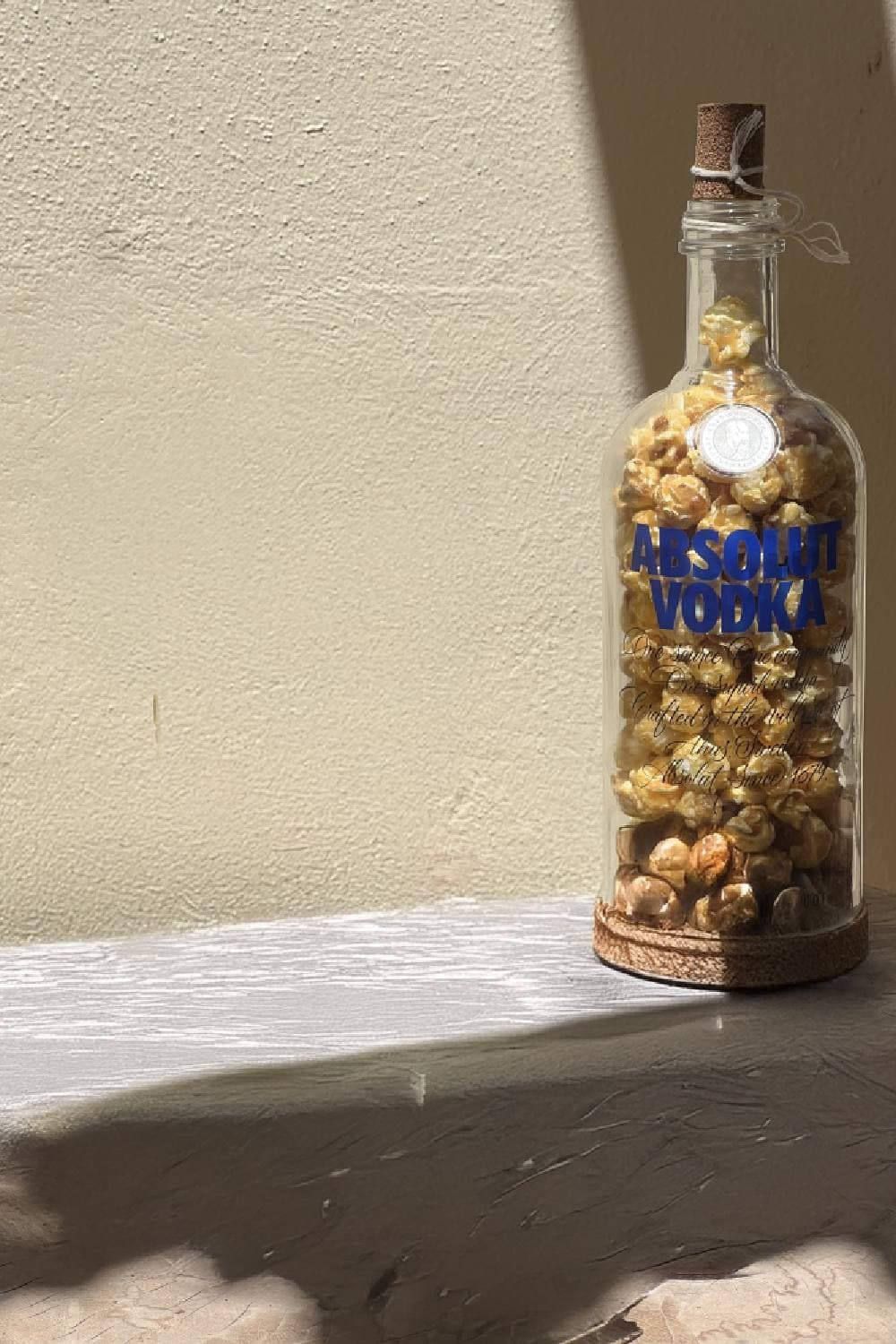 A glass bottle with a cork stopper tied on with twine, filled with golden popcorn and standing on a cork base in strong sunlight