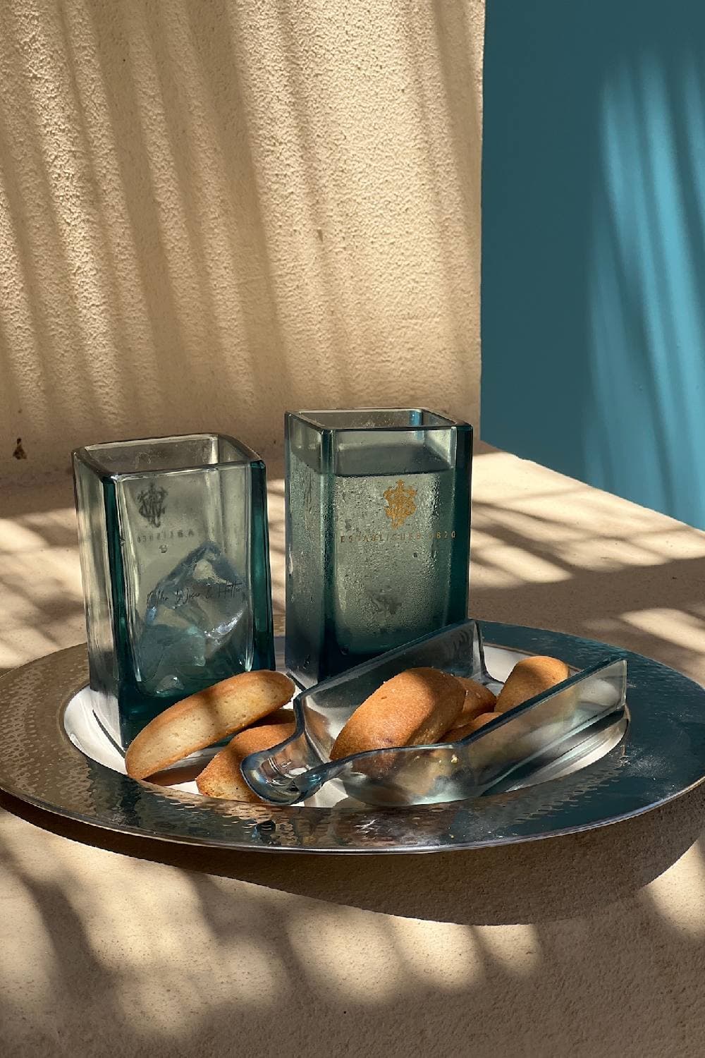 Two square glass tumblers and a bottle-shaped serving dish of small cakes on a hammered metal tray in dappled sunlight