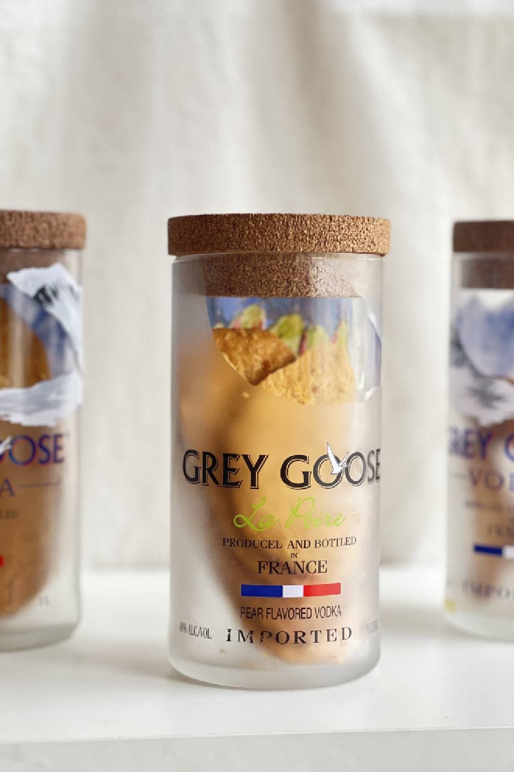 Frosted glass jars with cork lids on a white shelf, the middle one filled with snacks, against a linen backdrop