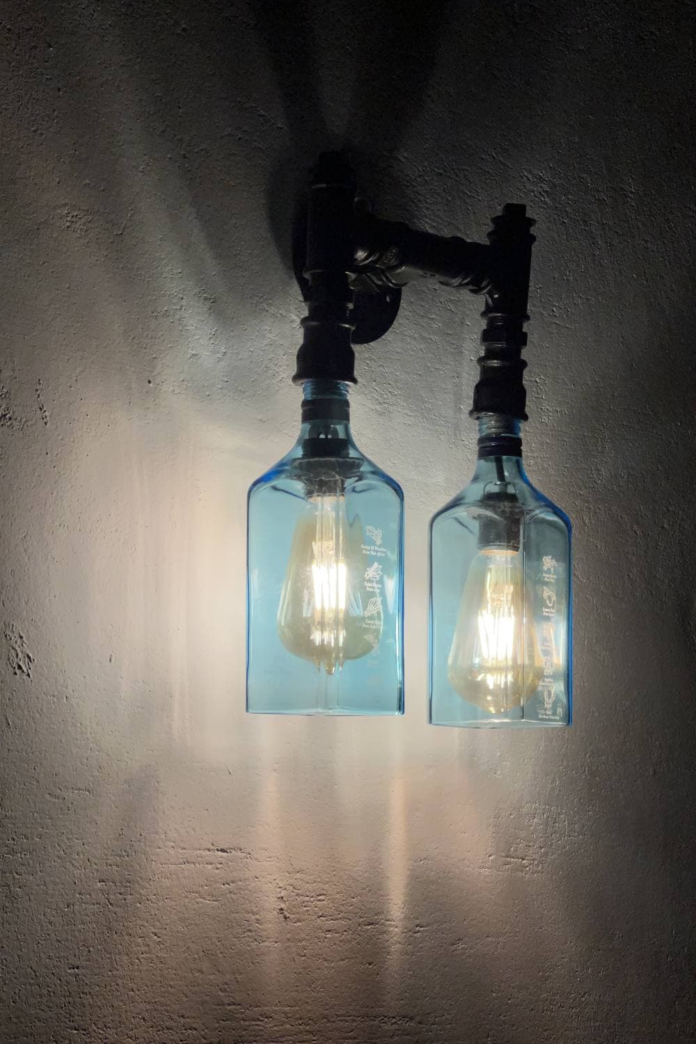Two blue glass bottle wall lights on black pipe fittings, glowing against a rough wall