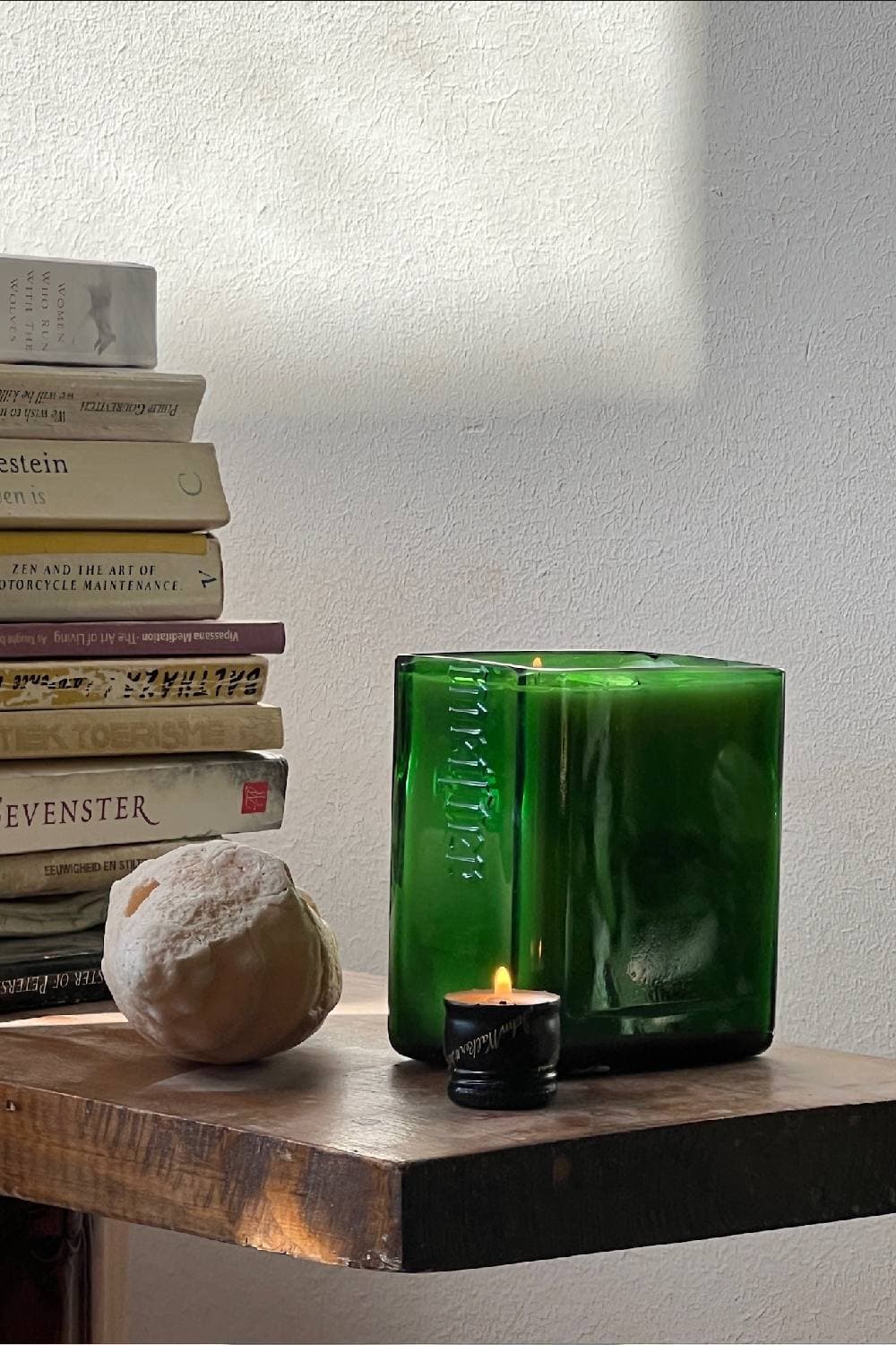 A square green glass candle beside a small black candle and a stack of books on a wooden table