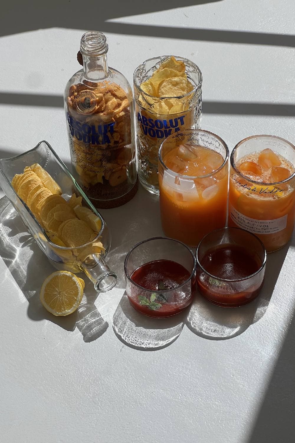 A table set from reused bottles: a corked bottle and a tall glass of snacks, two glasses of juice, two sauce bowls and a bottle-shaped chip dish