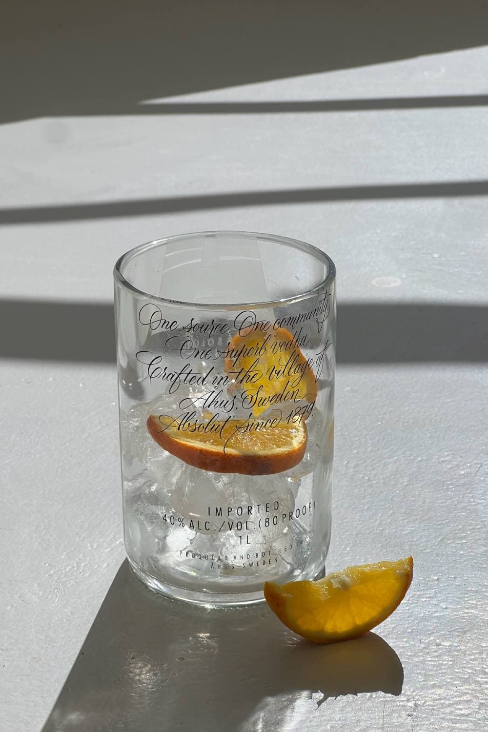 A clear tumbler printed with script lettering, holding ice and dried orange slices, with an orange wedge beside it